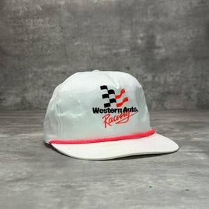 May include: White cap with a flat brim and a pink rope detail. The cap features the text "Western Auto. Racing" in black and red, along with a checkered flag and red wave design. A classic racing-themed accessory.