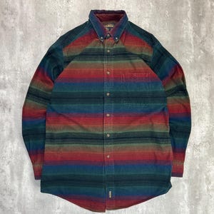 May include: A long-sleeved shirt with a striped pattern in red, green, blue, and brown. The shirt features a button-down collar and a single chest pocket. The fabric appears to be a soft, textured material. The shirt has a buttoned front.