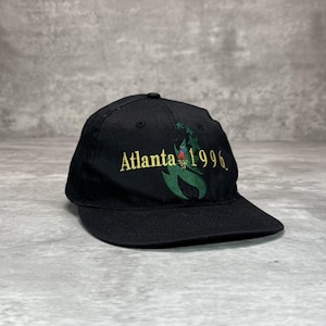以下が含まれることがあります： 金色の文字で「Atlanta 1996」と書かれた黒い野球帽。緑、赤、金色の刺繍デザインが施されています。つばはカーブしており、上部にボタンが付いています。