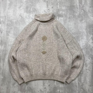 Op de afbeelding: Een beige coltrui met een geribbelde breistructuur en lange mouwen. Het midden van de borst is voorzien van een ronde geborduurde patch met tekst en daaronder een kleinere ruitvormige patch, beide in een bijpassende beige tint.