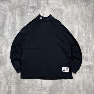 May include: Black long-sleeve shirt with a mock neck collar. The collar features the word "CHARGERS" and a team logo. A white tag with the word "MAXIT" is on the lower right. The shirt is made of a soft material.