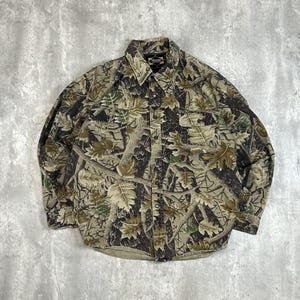 May include: A button-down camouflage shirt with a leaf pattern in shades of brown, green, and tan. The shirt has two chest pockets and a collar. The brand name "Master" is visible on the collar.