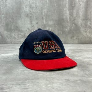 Può includere: Un cappellino da baseball blu navy con visiera rossa. La parte anteriore presenta un ricamo in oro e rosso con la scritta USA OLYMPIC TEAM. Una piccola toppa sul lato sinistro mostra il logo olimpico USA con il testo 100 e il marchio Champion.