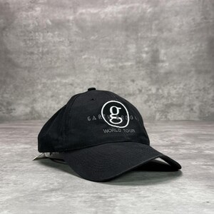Op de afbeelding: Zwarte baseballcap met een gebogen klep. De cap toont de tekst "GARTH BROOKS WORLD TOUR" in het wit, en een wit "g" logo in een cirkel.