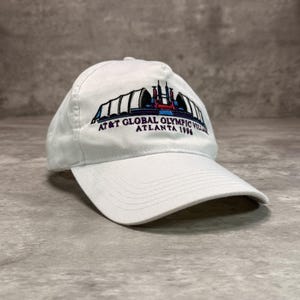 May include: A white baseball cap featuring an embroidered graphic of the AT&T Global Olympic Village Atlanta 1996. The design includes a stylized building structure in blue, red, and black thread centered on the front panel above the brim.