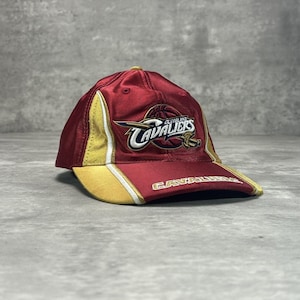 May include: A maroon and gold baseball cap featuring the embroidered Cleveland Cavaliers logo on the front crown. The cap includes white piping accents along the side panels and the word Cavaliers stitched in gold on the curved brim.