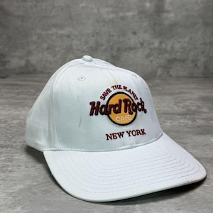 Op de afbeelding: Een witte baseballpet met een geborduurd Hard Rock Cafe logo op de voorzijde. Het logo bevat een gele cirkel met de tekst Hard Rock Cafe in donkerrood, omringd door de woorden Save The Planet en New York in het rood.