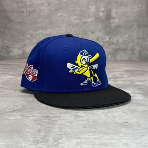 Peut inclure: Casquette de baseball bleue avec une visière noire. La casquette présente un personnage de dessin animé tenant une batte de baseball et les mots "West League" sur le côté. Le chapeau a un bouton noir sur le dessus.