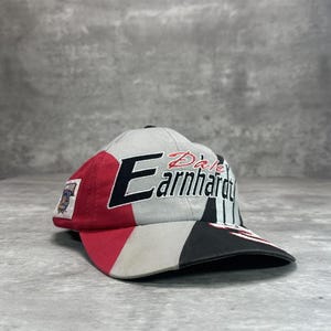 Op de afbeelding: Een veelkleurige baseballcap met de naam "Dale Earnhardt" in rode en zwarte letters. De cap heeft een kleurenschema van rood, zwart, grijs en wit. Een patch is zichtbaar aan de zijkant.