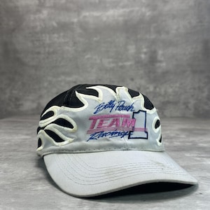 May include: A gray baseball cap with a black top and a curved brim. The cap features a flame design and the text "Billy Roach TEAM Racing" in pink and blue. The number "1" is also visible.