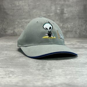 May include: A grey baseball cap featuring an embroidered graphic of a cartoon grim reaper character standing on a yellow patch of ground. The word Blind is embroidered in white next to the character, and the brim has a dark blue trim.