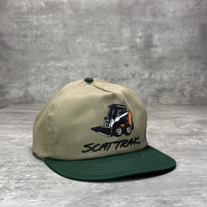 Può includere: Un cappellino da baseball color marrone chiaro con visiera verde scuro e un bottone verde scuro sulla parte superiore. La parte anteriore presenta un grafico ricamato di una minipala bianca e nera con accenti arancioni e il testo nero SCAT TRAK.