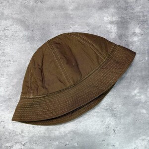 May include: A solid brown bucket hat featuring a rounded crown and a downward-sloping brim. The hat is constructed from a smooth fabric with visible tonal stitching details along the seams of the crown and multiple rows of stitching on the brim.