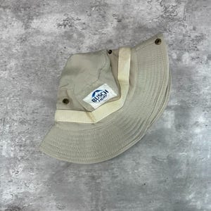 May include: A tan bucket hat with a white strap and the "Busch Light" logo. The hat is made of a light fabric and has a wide brim. The hat is a neutral color and is suitable for outdoor activities.