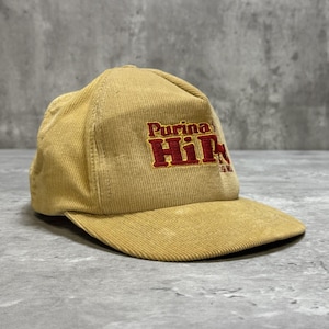 Op de afbeelding: Een beige corduroy baseballpet met de geborduurde tekst Purina HiPro in rode en gele letters op het voorpaneel. De pet heeft een gestructureerde kroon, een stijve klep en een textuur van stof over het gehele buitenoppervlak.