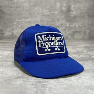 May include: A blue trucker hat with a mesh back and a curved brim. The front panel features the text "Michigan Propellers" in white, within a white rectangular border. The hat is a vibrant blue color.