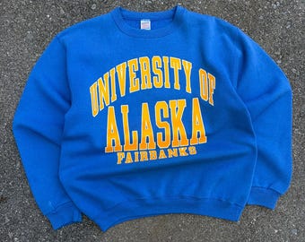 May include: A bright blue crewneck sweatshirt featuring the text UNIVERSITY OF ALASKA FAIRBANKS printed in bold yellow block letters across the chest. The garment has long sleeves, a ribbed collar, and ribbed cuffs, with a small white tag at the neck.