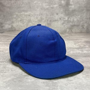 Op de afbeelding: Een effen koningsblauwe baseballpet met een gestructureerde kroon van zes panelen, een platte klep en een kleine met stof beklede knoop in het midden bovenop. De pet is voorzien van geborduurde oogjes op elk paneel voor ventilatie.