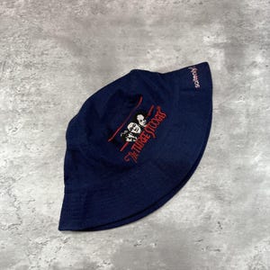 May include: A navy blue bucket hat featuring an embroidered design of The Three Stooges in red and white. The hat has a soft, unstructured shape and a small brim. The word "soitenly!" is embroidered on the side.