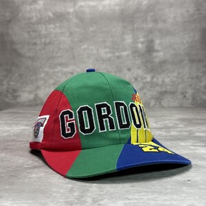 Op de afbeelding: Een kleurrijke baseballcap met de naam "GORDON" in grote, zwarte letters. De cap heeft een patchwork-ontwerp met rode, groene en blauwe panelen. Extra tekst en logo's zijn zichtbaar op de zijkant en de klep.