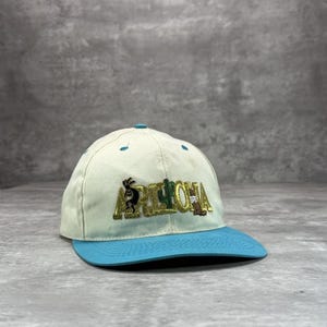 May include: A cream-colored baseball cap with a turquoise brim. The cap features the word "ARIZONA" embroidered in gold, along with desert-themed graphics. The cap has a curved brim and a button on top.