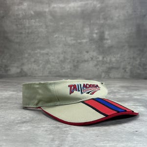 Peut inclure: Visière beige avec le mot "TALLADEGA" en rouge et bleu. La visière présente un motif rayé rouge, bleu et blanc sur le bord. La visière est faite d'un matériau semblable à de la toile.