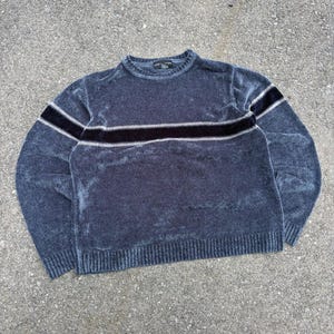 Può includere: Un maglione a maniche lunghe con girocollo in tessuto grigio antracite scuro dall'aspetto vellutato. Il capo presenta una striscia orizzontale nera sul petto, delimitata da sottili linee bianche. I polsini, lo scollo e l'orlo sono a coste.