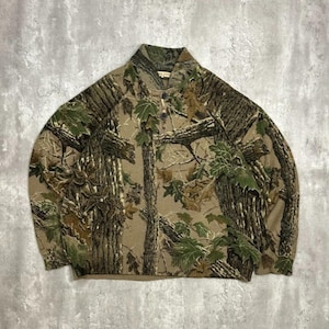 May include: A long-sleeved shirt with a camouflage design, showing brown tree trunks and green leaves. It has a collared neckline and three buttons. The colour scheme is earthy, ideal for outdoor pursuits.