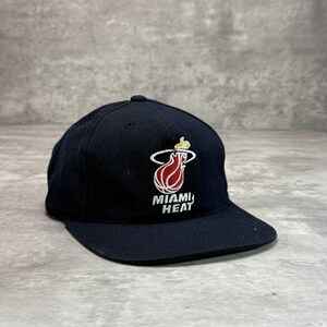 Op de afbeelding: Zwarte baseballcap met het Miami Heat-logo geborduurd op de voorkant. Het logo is een gestileerde vlam in rood, geel en wit, met de woorden "MIAMI HEAT" eronder. De cap heeft een gebogen klep.