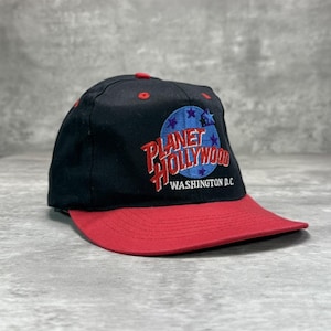 Op de afbeelding: Zwarte en rode Planet Hollywood baseball cap. De cap heeft het Planet Hollywood logo in rood en wit met een blauwe achtergrond en sterren. De tekst "WASHINGTON D.C." is ook zichtbaar.