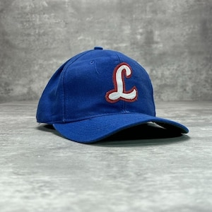 Op de afbeelding: Een blauwe baseballpet met een gebogen klep. De pet heeft een grote, gestileerde letter "L" op de voorkant, omlijnd in wit met een rode binnenkant. De pet is gemaakt van een zachte stof en heeft een knoop aan de bovenkant.