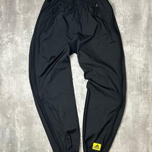 May include: Black athletic trousers with a drawstring waist and zipped pockets. The trousers have zips at the ankles and a yellow Adidas logo patch. The material appears to be a lightweight nylon.