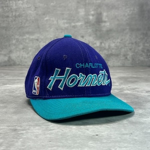 May include: A Charlotte Hornets baseball cap in a classic style. The cap features a royal blue crown and a teal green bill. The team name "Hornets" is embroidered in teal, with "Charlotte" above it. The NBA logo is on the side.