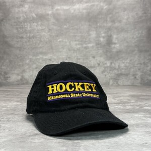 May include: Black baseball cap featuring the word "HOCKEY" in gold lettering above "Minnesota State University" in white. The hat has a curved peak and is made of a soft fabric.