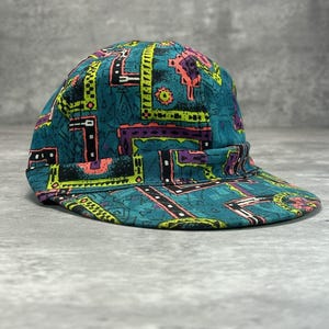 May include: A teal and patterned cap with a short brim. The cap features a geometric design in neon green, pink, purple, and black. The cap is made of a durable fabric and is suitable for outdoor activities.