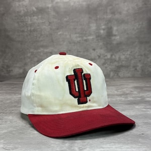 Op de afbeelding: Een witte en rode baseballcap met het Indiana University logo in rood. De cap heeft een rode klep en een witte kroon met rode accenten. Het logo is op de voorkant geborduurd.