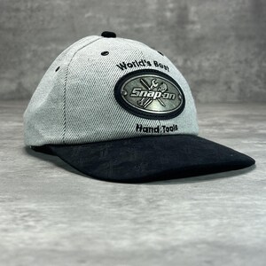 May include: A grey and black baseball cap featuring the text "World's Best" and "Snap-on Hand Tools". The cap has a curved peak and a black button. The Snap-on logo is in a silver oval on the front.