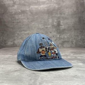 Op de afbeelding: Een blauwe denim baseball cap met geborduurde cartoonfiguren. De cap heeft een gebogen klep en een ronde kroon. Het geborduurde ontwerp bevat personages als Taz en Bugs Bunny. De cap is lichtblauw.