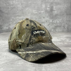 May include: A baseball cap featuring a camouflage pattern in shades of brown, tan, and green. The front of the cap displays a black rectangular patch with the white embroidered text "DAYTON FREIGHT" centered on the crown above the curved brim.