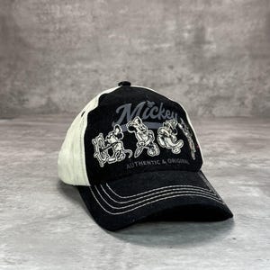 May include: Black and white baseball cap featuring Mickey Mouse. The cap has a black brim and crown with a white mesh back. The word "Mickey" is printed above three Mickey Mouse illustrations. The text "AUTHENTIC & ORIGINAL" is below the illustrations.