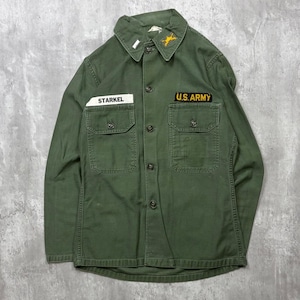May include: An olive green long-sleeve button-up shirt featuring two chest pockets, a collar, and embroidered patches. The right chest has a white name tag reading STARKEL, while the left chest has a black and yellow U.S. ARMY patch.