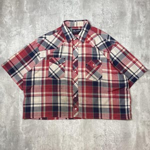 May include: A short-sleeved, button-down shirt in a plaid pattern of red, white, and navy blue. The shirt has two chest pockets and a collar. The brand name "Wrangler" is visible on the collar.