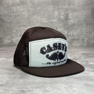 May include: A brown trucker hat featuring a white front panel with a black graphic of a mustache and the text "CASEY'S ...the original roadhouse". The hat has a solid brown brim and a mesh back panel, constructed with a structured crown and a snapback closure.