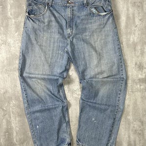 May include: Light blue denim jeans with a classic five-pocket design. The jeans show signs of wear, with faded areas and slight distressing. The relaxed fit and light wash give them a vintage look.