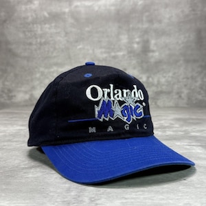 May include: A black and blue baseball cap featuring the text "Orlando Magic" in white and blue. The cap has a blue peak and a blue button on top. A classic style hat.