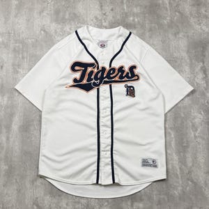 May include: A white short-sleeved baseball jersey featuring the word Tigers in navy blue with orange trim across the chest. The shirt has a navy blue vertical stripe along the button placket, a small navy D logo, and a rectangular MLB patch at the hem.