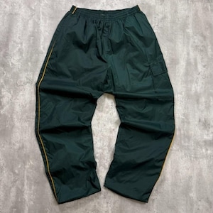 May include: Dark green nylon athletic trousers with a cargo pocket on the right leg. The trousers have a yellow stripe down the side seams and along the top waistband. The trousers are laid flat on a grey surface.