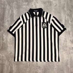 May include: Black and white striped polo shirt with a black collar and a quarter-zip closure. The shirt features a small emblem on the chest and short sleeves. The shirt is a classic design.