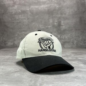 Op de afbeelding: Een baseballpet met een witte kroon en een zwarte klep. De pet heeft het woord "Hawaii" boven een handgebaar en de woorden "HANG LOOSE" in zwarte letters.