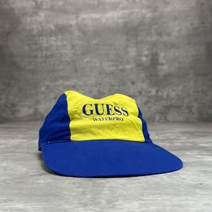 Könnte beinhalten: Ein blau-gelbes Guess Waterpro Visier. Das Visier hat einen gebogenen Schirm und ein gelbes Oberteil mit dem Wort "GUESS" und "WATERPRO" in Blau aufgedruckt. Der Rest des Visiers ist blau.